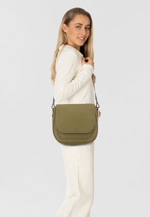Donna con lunghi capelli biondi, vestita con un outfit bianco, mentre porta una borsa a tracolla di colore verde oliva con patta sulla spalla, sorridendo leggermente.