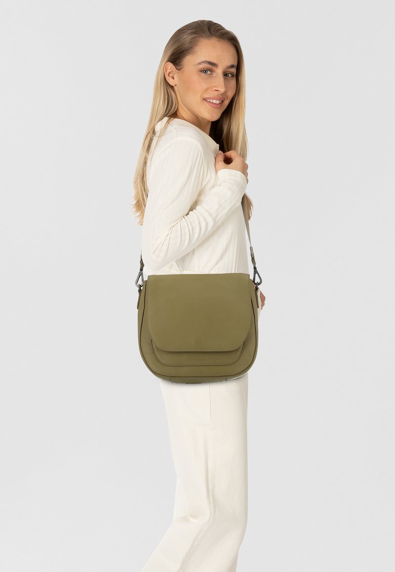 Donna con lunghi capelli biondi, vestita con un outfit bianco, mentre porta una borsa a tracolla di colore verde oliva con patta sulla spalla, sorridendo leggermente.