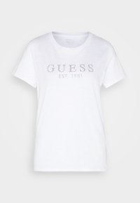 Bílý bavlněný tričko s kulatým výstřihem, krátkými rukávy a zdobeným logem "GUESS EST. 1981" na přední straně. Hladká textura.