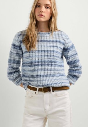 Jeune femme aux longs cheveux blonds portant un pull en mailles rayé bleu et blanc, rentré dans un pantalon blanc avec une ceinture marron.
