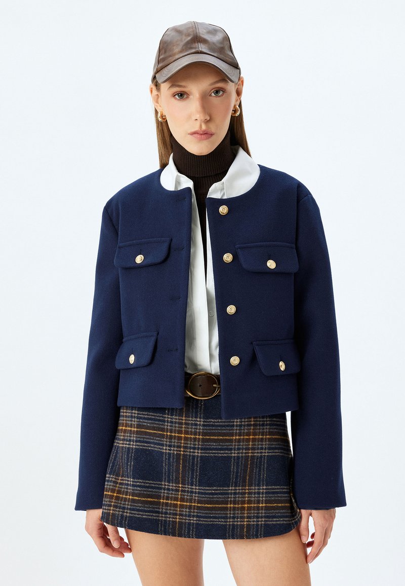 Giacca in misto lana blu navy con colletto rotondo, bottoni dorati e due tasche frontali, abbinata a una gonna a quadri e un berretto marrone.