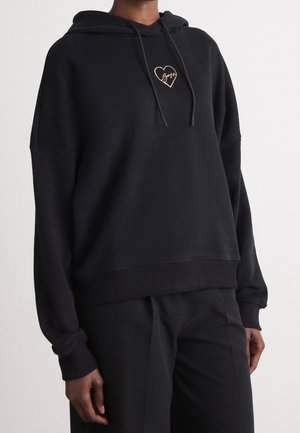 Person wearing a black hoodie with drawstrings and a small rose gold heart emblem on the chest, paired with black pants.