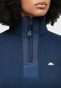 Marineblå fleecegenser med høy krage, halvglidelås og teksturert stoff. Har en liten hvit logo på brystet.