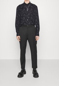 Zwart overhemd met paisley-patroon, gecombineerd met hooggetailleerde kolenstofbroek en zwarte schoenen met dikke zolen. Slim fit met knoopsluiting.