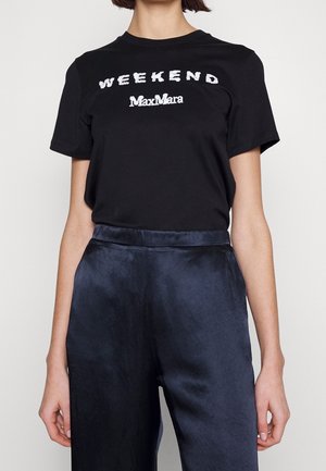 Person iført en sort t-shirt med teksten "WEEKEND MaxMara" og marineblå satinbukser, stående mod en ensfarvet lys baggrund.