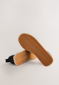 Black slip-on shoes with a textured surface, featuring a woven jute detail and a rubber outsole with a herringbone pattern.