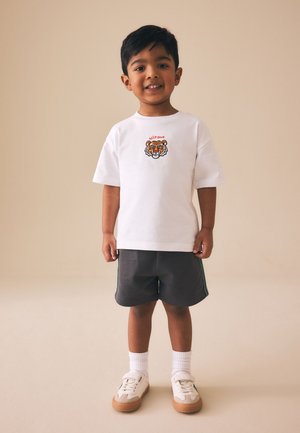 Fiatal fiú áll és mosolyog, fehér pólót visel tigris grafikával, sötét rövidnadrágot, fehér zoknit és fehér tornacipőt.