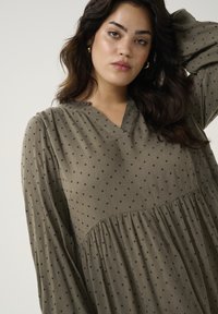 Robe vert olive avec des pois noir, décolleté en V, manches longues, corsage froncé et tissu texturé. L'ourlet s'évase doucement.