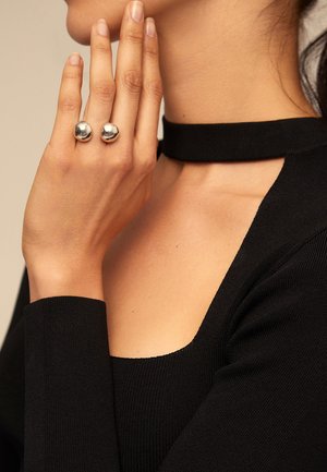 Top negro acanalado con cuello alto, mostrando una mano que lleva dos anillos plateados brillantes y esféricos en los dedos. Detalles del tejido texturizado visibles.