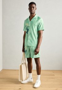 Kortærmet grøn skjorte og matchende shorts i tie-dye mønster med hvide blomster grafik. Suppleret med en beige tote bag og hvide sneakers.