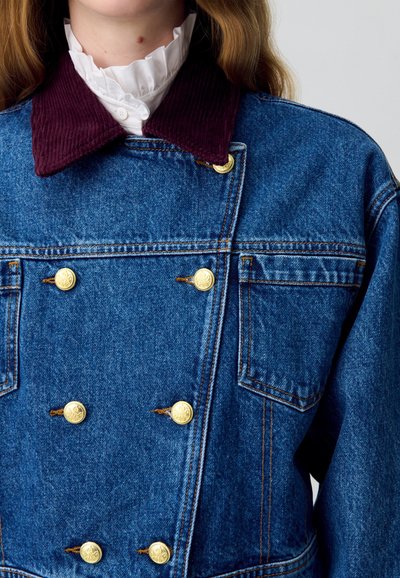 Giacca di denim con un colletto in corduroy color bordeaux scuro, dettagli con bottoni dorati e cuciture blu a contrasto, che incarna una silhouette strutturata.