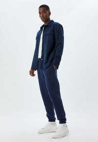 Camisa de botones azul marino con dos bolsillos en el pecho, combinada con pantalones jogger a juego que tienen cremalleras laterales y zapatillas blancas.