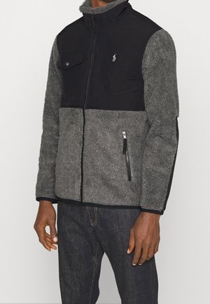 Homme portant une veste polaire zippée bicolore grise et noire avec une poche poitrine et des poches latérales zippées.