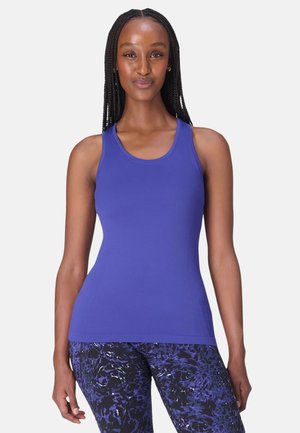 Vrouw met gevlochten haar, een nauwsluitende blauwe mouwloze top en zwart-blauwe patroonleggings, staand tegen een effen achtergrond.