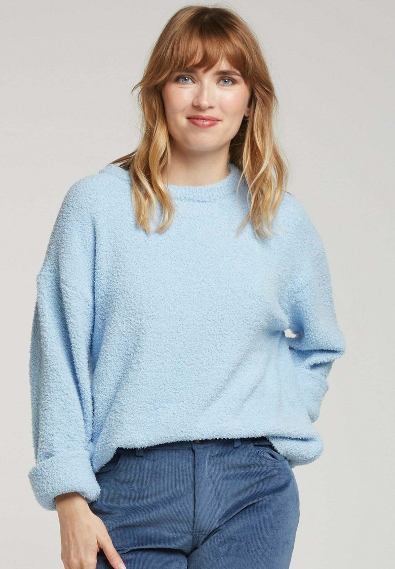 Un pull-over léger bleu clair avec une texture duveteuse, un col rond et des épaules tombantes, associé à un pantalon en velours côtelé bleu foncé.