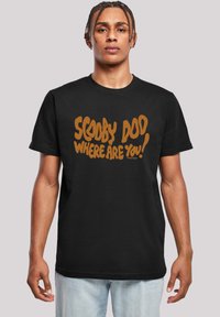 Černé bavlněné tričko s rezavě hnědým textem: „SCOOBY DOO, KDE JSI!“ Design má tučný, hravý font. Pohodlný střih s krátkým rukávem.