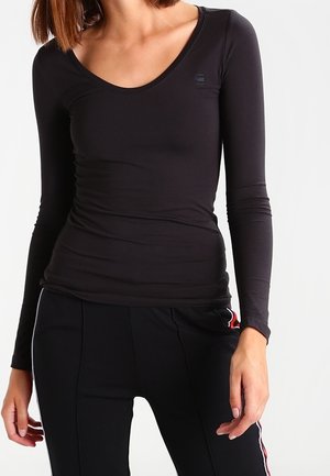 Mujer que lleva una camiseta negra ajustada de manga larga con escote redondo y pantalones negros con franjas laterales rojas y blancas.