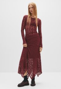 Burgunderfarbenes Spitzenkleid mit langen Ärmeln, tailliertem Oberteil und asymmetrischem Saum. Kombiniert mit dunklen Socken und Slippern, die eine kunstvolle Textur zeigen.