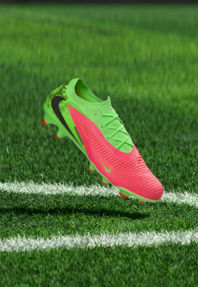 Φωτεινό πράσινο και ροζ ποδοσφαιρικό παπούτσι με μαύρο σήμα, αιωρούμενο πάνω από τεχνητό γρασίδι με λευκές γραμμές.