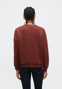 Bordeauxrød sweatshirt i bomuldsblanding med rund halsudskæring, sænkede skuldre, ribstrikkede manchetter og kant. Brugt med mørke jeans, set fra bagsiden.