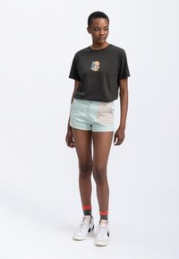 Zwarte t-shirt met een grafisch ontwerp, lichtgroene shorts met een beige patch, en witte sneakers met rode en grijze accenten.