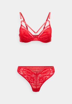 Soutien-gorge en dentelle rouge avec des bretelles croisées et des bonnets triangulaires. String en dentelle rouge assorti avec un motif floral et des accents. Tissu doux et transparent.