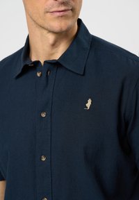 Polo kratkih rukava u tamnoplavoj boji od lagane tkanine, sa dugmadi sprijeda i malim bež izvezom logom na lijevoj strani prsiju.