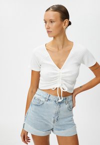 Witte geribbelde crop top met een diepe V-hals en een gatherings detail met trekkoord aan de voorkant, gecombineerd met lichtblauwe denim shorts.