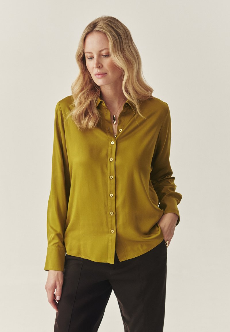 Olivegrünes Blusenshirt mit langen Ärmeln aus glattem Stoff, mit einer durchgehenden Knopfleiste aus goldfarbenen Knöpfen und einem klassischen Kragen. Schwarze Hosen sichtbar.