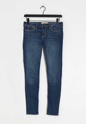 Jeans slim bleu foncé suspendu sur un cintre noir sur fond blanc, montrant les poches avant et la fermeture par boutons.