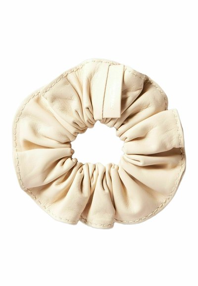 Scrunchie in pelle beige con design di tessuto arricciato e cuciture visibili. Texture liscia, forma circolare, con un piccolo etichetta attaccata.