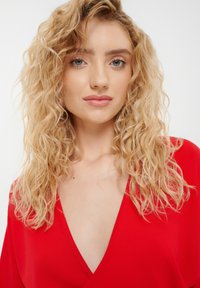 Červený top s hlbokým V-výstrihom a splývavými rukávmi; textúrovaná tkanina; jemné, vlnité blond vlasy a prirodzený make-up. Jednoduché pozadie.