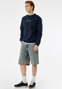 Sudadera navy con cuello redondo y texto en la parte frontal, combinada con pantalones cortos de mezclilla azul claro. El modelo lleva calcetines blancos y zapatillas negras.