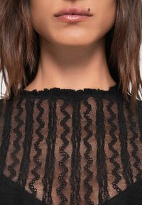 Top in pizzo nero con scollatura a onde, motivo zigzag e design testurizzato. Il tessuto è trasparente e presenta delicati dettagli in tutto il capo.