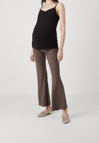 Gravid kvinna i svart ärmlös topp, bruna utsvängda byxor och guldvävda flats som står mot en vit bakgrund.