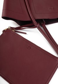 Burgundska usnjena torba s ravnimi ročaji, ki vključuje usklajen žep za zadrgo. Gladka tekstura, minimalističen dizajn, izbočen logotip.