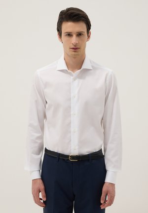 Mann trägt ein weißes Hemd mit Knöpfen, in dunkelblaue Hose mit schwarzem Gürtel gesteckt, vor einem schlichten hellen Hintergrund.