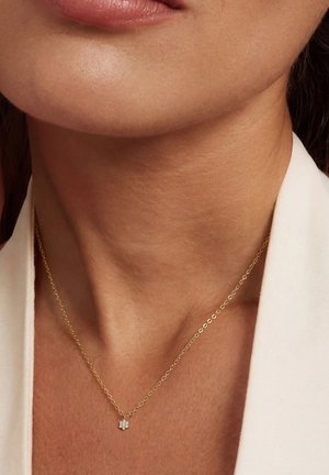 Close-up of neck with a delicate gold chain necklace featuring a small flower-shaped diamond pendant, worn with a white top.