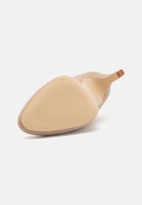 Scarpa stiletto di camoscio beige singola, mostrata con la suola rivolta verso l'alto, con una texture antisdrucciolevole nella parte anteriore della suola su uno sfondo bianco.