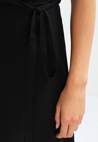 Černá látka šatů s jemnou texturou, s uvázaným páskem v pase. Detailní záběr ukazuje ruku držící pásek.
