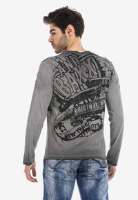 Graues Langarmshirt mit verblasster Textur, ausgestattet mit schwarzem grafischem Text und einem Design, das einem Vintage-Emblem auf der Rückseite ähnelt.