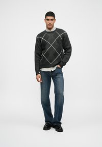 Szary sweter w wzór w diamenty, założony na jasną koszulę z kołnierzykiem, w połączeniu z niebieskimi dżinsami i czarnymi butami.