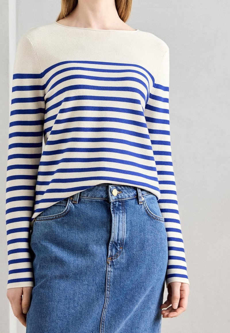 Femme portant un haut à manches longues crème et bleu à rayures horizontales, rentré dans une jupe en denim bleu taille haute avec une taille boutonnée.