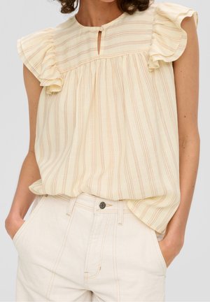 Kvinde iført en cremetærmet bluse uden ærmer med subtile lodrette striber og flæser på skuldrene, kombineret med cremefarvede, taljehøje bukser med lommer.