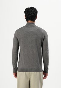 Uomo che indossa un maglione a collo alto grigio scuro e pantaloni beige, in piedi con la schiena rivolta verso la fotocamera su uno sfondo bianco semplice.
