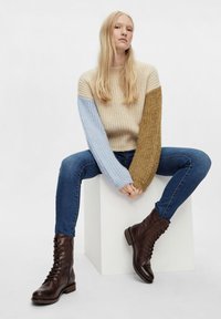 Suéter de punto con cuerpo beige, mangas azul claro y marrón dorado; jeans azules; botas de cuero marrón con cordones; sentado en un cubo blanco.