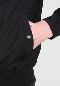 Czarna nylonowa kurtka z prążkowanymi dzianinowymi mankietami i pasem. Posiada boczną kieszeń z okrągłym logo. Gładka faktura, dopasowany fason.