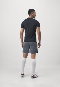 Svart idrottströja med korta ärmar, grå shorts, vita knästrumpor och fotbollsskor med röda accenter, sedd från baksidan.