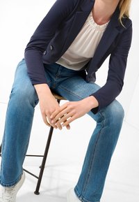 Granatowa marynarka na białej bluzce z falbankami w połączeniu z jasnoniebieskimi jeansami o prostym kroju i białymi trampkami, siedząca na metalowym stołku.