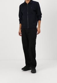 Sudadera azul marino con cremallera y cuello, combinada con pantalones negros de corte holgado y zapatillas negras. Diseño sencillo, sin patrones.